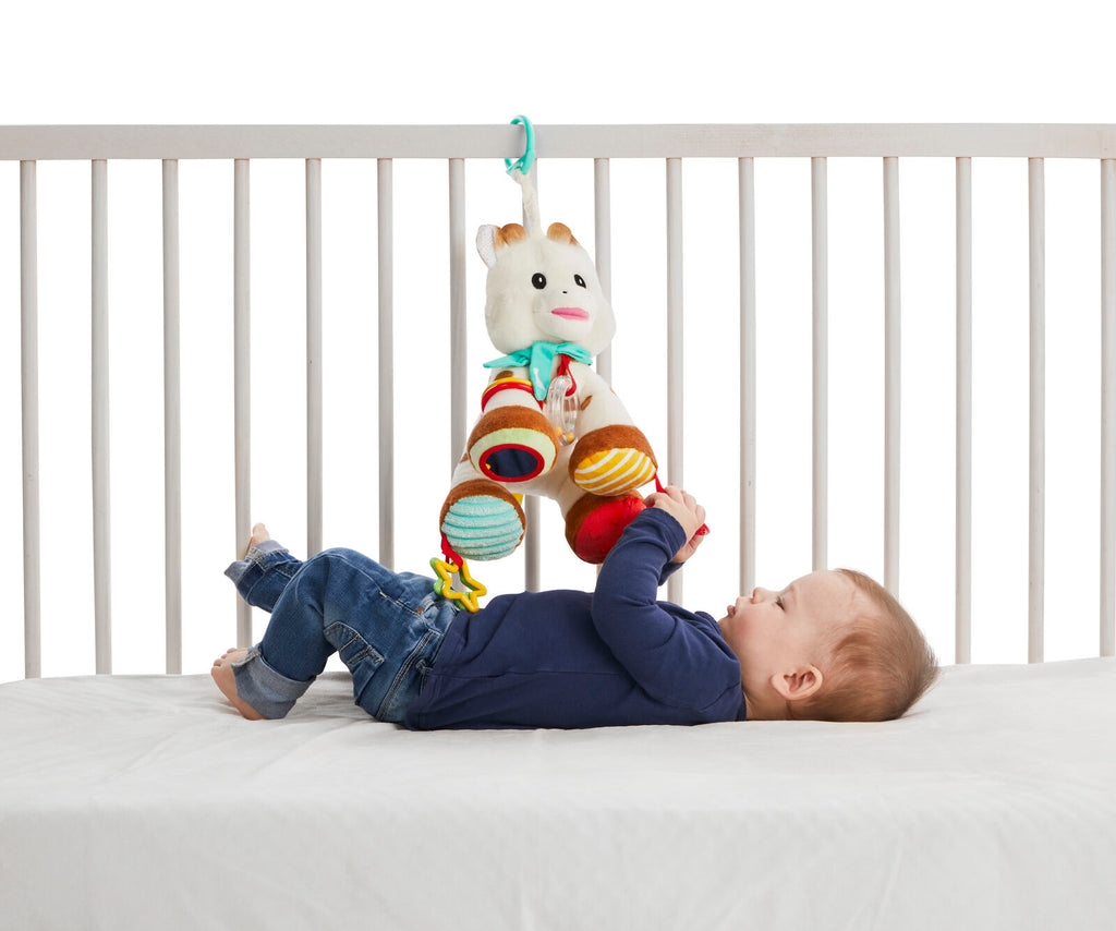 Sophie activities plush hanging from crib while toddler plays